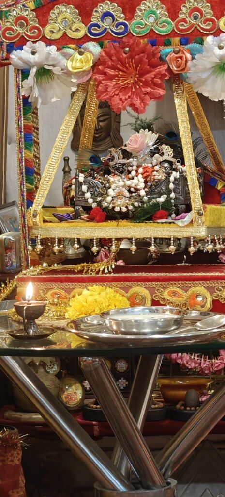Laddu Gopal in a flower-decorated jhula with a lit diya and bhog for Janmashtami